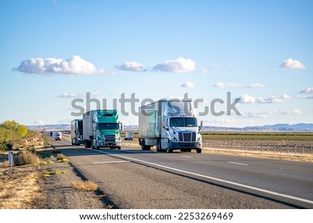 Industrial commercial big rigs long hauler semi trucks with semi trailers transporting different cargo driving on the wide divided straight highway road between the fields in California Royalty-Free Stock Photo #2253269469
