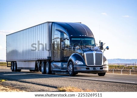 Industrial long hauler big rig black semi truck tractor with truck driver sleeping compartment transporting cargo in dry van semi trailer driving on the straight wide highway road in California Royalty-Free Stock Photo #2253269511