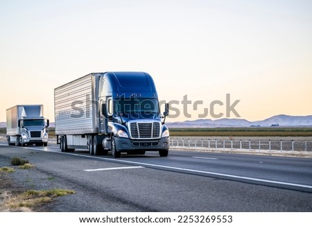 Industrial commercial big rigs long hauler semi trucks with semi trailers transporting different cargo driving on the wide divided straight highway road between the fields in California Royalty-Free Stock Photo #2253269553