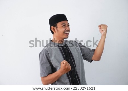 Successful happy young asian muslim man screaming shouting and shows winning victory gesture wearing grey muslim clothes isolated on white background