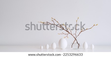 Minimalistic Easter still life. A glass vase with a branch on which colorful Easter eggs hang on a white background. Interior photography. It's springtime.