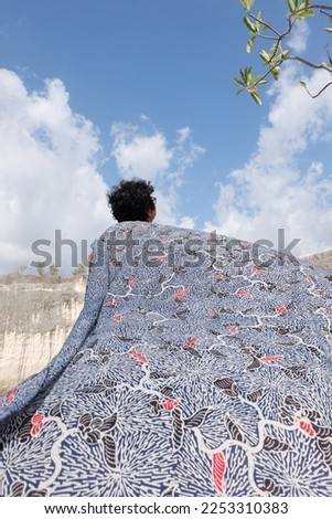 A girl holding and waving a blue Javanese batik fabric (also called batik tenun jawa) with sky background