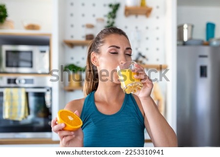 Beautiful young woman drinking fresh orange juice in kitchen. Healthy diet. Happy young woman with glass of juice and orange at table in kitchen.