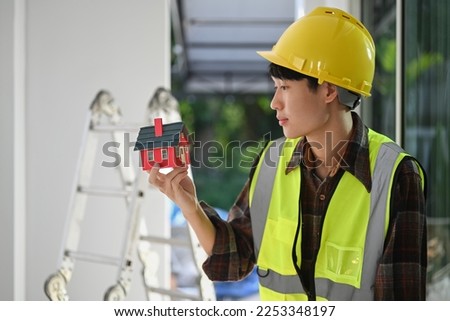 Photo of smiling construction worker in safety helmet and vest holding house model