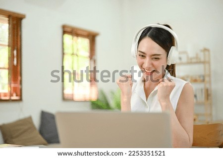 Cheerful and attractive millennial Asian woman wearing headphones, looking at laptop screen, receiving a good unexpected email, celebrating her success.