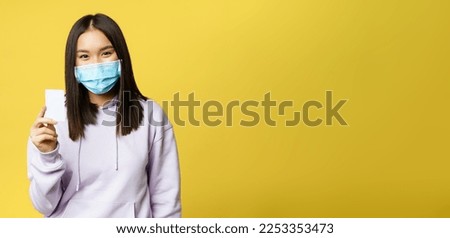 Asian woman in medical face mask, showing credit card, pass, standing over yellow background.