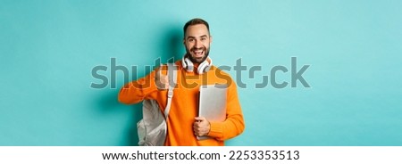 Happy man with backpack and headphones, holding laptop and smiling, showing thumb-up in approval, standing over turquoise background.