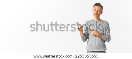 Image of thoughtful middle-aged man making choice, looking and pointing left, standing over white background.