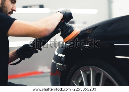 Caucasian man wearing black gloves crouching by the front of a black car polishing it using professional tool. Car detailing process. Horizontal indoor shot. High quality photo