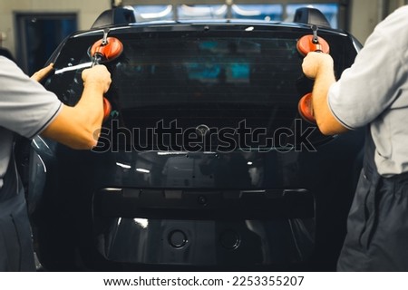 Rear view of two men installing rear window pane into back of car using professional tools. Garage work. Car maintenance. Horizontal indoor shot. High quality photo