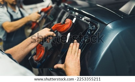 Close-up of two unrecognisable men holding rear window pane to install into back of car. Car maintenance. Garage work. Horizontal indoor shot. High quality photo Royalty-Free Stock Photo #2253355211
