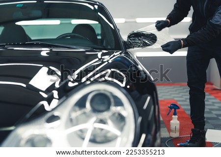 Unrecognisable man wearing black gloves stretching PPF Paint Protection Film over side mirror of black car. Professional car detailing. Horizontal indoor shot. High quality photo