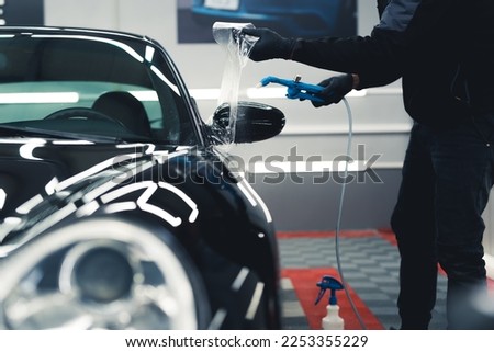 Man wearing black gloves and work uniform applying PPF Paint Protection Film to side mirror of black car. Car detailing process. Horizontal indoor shot. High quality photo