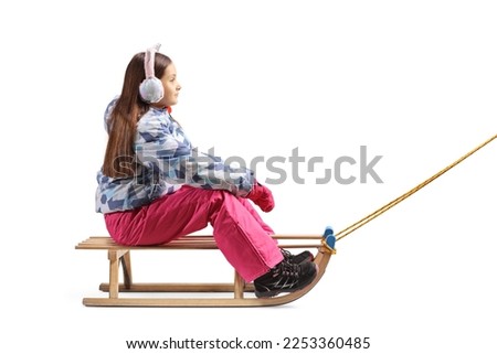 Full length profile shot of a girl with earmuffs sitting on a wooden sleigh isolated on white background