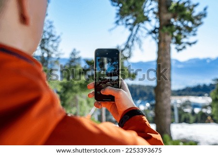 A teenage boy dressed in ski clothes, taking a picture of the snowy landscape with his mobile phone in his hand, during the winter season.