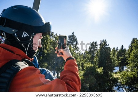 A teenage boy dressed in ski clothes, taking a picture of the landscape with his mobile phone in his hand, while sitting on a ski lift that takes him to the ski slopes, during the winter season.