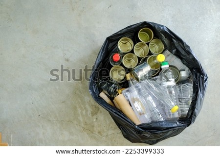 Top view of unsorted plastic glass bottles metal cans containers paper cardboard collected in black trash bag on cement floor. Zero waste sustainability concept. Selective focus. Copy space on left Royalty-Free Stock Photo #2253399333