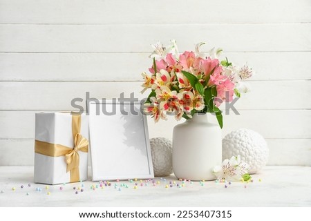 Vase with beautiful alstroemeria flowers, empty picture frame, gift and decor on table near light wooden wall. Mother's day celebration