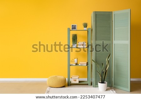 Interior of room with folding screen, shelving unit and houseplant near yellow wall Royalty-Free Stock Photo #2253407477