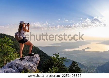 A happy contented young girl with a backpack takes a souvenir photo of the Montenegrin coastal cities, the Adriatic Sea and the bright sunset rays from the top of a high mountain
