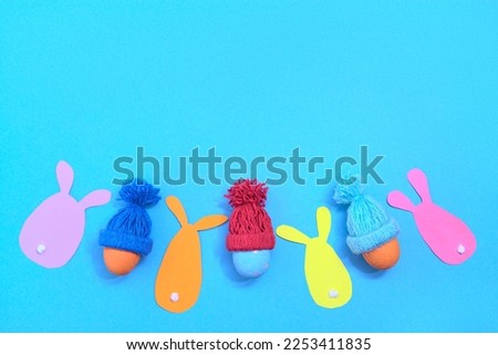 Painted Easter eggs in a winter knitted hat. Rabbit cut out of coloured paper. Blue background. Copy scape.