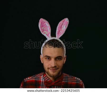 Caucasian man with pink Easter bunny ears. Portrait European in red shirt in dark green studio. Emotion of cunning. Man has tricky evil plan and looks mysteriously at camera, think over good idea.