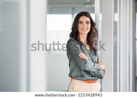Portrait of mature business woman with arms crossed looking at camera. Happy smiling mid businesswoman standing in office with copy space. Confident latin woman in formalwear standing in workplace. Royalty-Free Stock Photo #2253428943