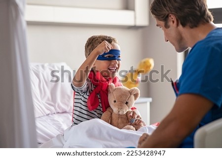 Strong little boy wearing superhero blue mask and red cape sitting on hospital bed playing with nurse. Happy superhero kid gesturing with playful doctor at clinic. Happy child playing as superman.