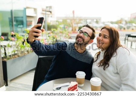 Cheerful influencer boyfriend and chubby girlfriend smiling taking a selfie with a smartphone while at the coffee shop to post it together on social media 