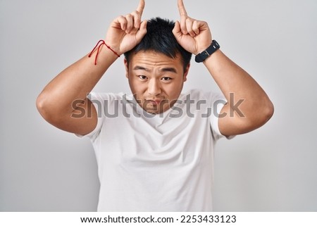 Young chinese man standing over white background doing funny gesture with finger over head as bull horns 