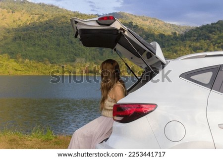 Woman traveler sitting on hatchback car on vacation with river mountain in background.
Freedom road trip journey concept.