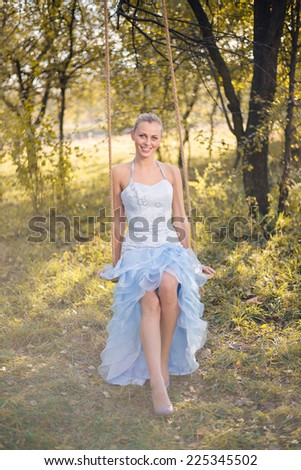 Beautiful young woman in prom dress sitting on swings on green summer or autumn outdoors copyspace background