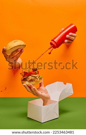 Food pop art photography. Female hand sticking out orange paper and pouring ketchup on burger on hand sticking out food box. Taste, creativity, art. Complementary colors. Copy space for ad, text