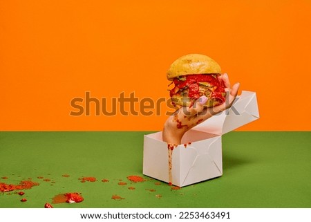 Food pop art photography. Female hand sticking out food box with burger on green tablecloth and orange background. Concept of taste, creativity, art. Complementary colors. Copy space for ad, text