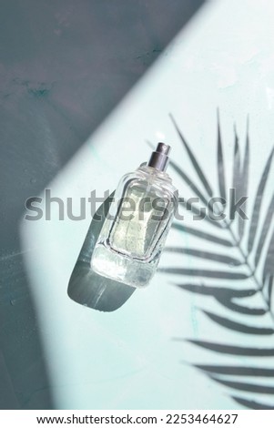 Glass blue transparent bottle of perfume is laying on the blue background with palm leaves shadow. Sunshine shade and sparkles. Tropical summer. Hawaii island.