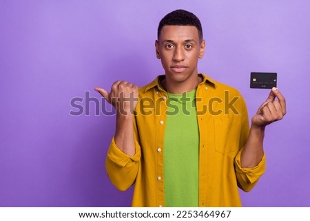 Photo portrait of nice young guy hold credit card point empty space eshop promo wear trendy yellow look isolated on purple color background