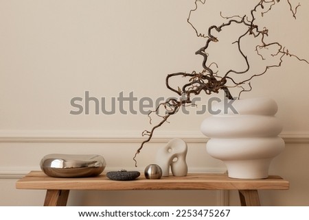 Warm and cozy composition of living room interior with wooden bench, modern beige vase with branch, silver sculpture, beige wall with stucco and personal accessories. Home decor. Template. Royalty-Free Stock Photo #2253475267
