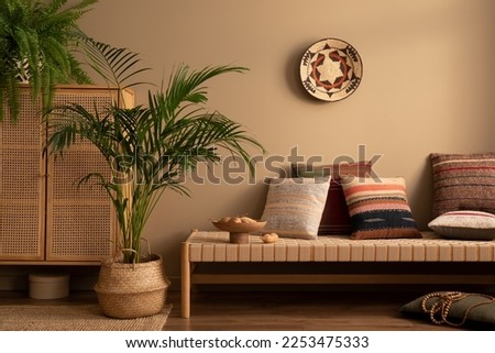 Warm and cozy ethno living room interior with couch, patterned pillows, plants i flowerpots, fern, rattan sideboard, basket on wall, wooden bowl and personal accessories. Home decor. Template. Royalty-Free Stock Photo #2253475333