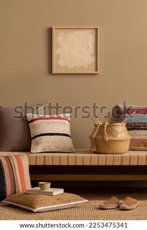 Interior design of ethno living room interior with mock up poster frame, colorful pillows, braided basket, rug, beige book, cup, brown wall, wooden floor and personal accessories. Home decor. Template Royalty-Free Stock Photo #2253475341