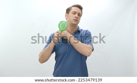 Young man isolated on white background cooling off on a hot day with a small fan.Overheated young man suffering from high temperature summer heat problem. Royalty-Free Stock Photo #2253479139