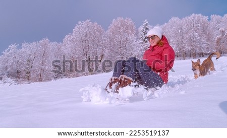 CLOSE UP: Cheerful woman, followed by cute dog, is sledding down a snowy hill. Young and smiling lady riding sled in fresh snow and enjoying outside on a gorgeous winter day with her furry friend.