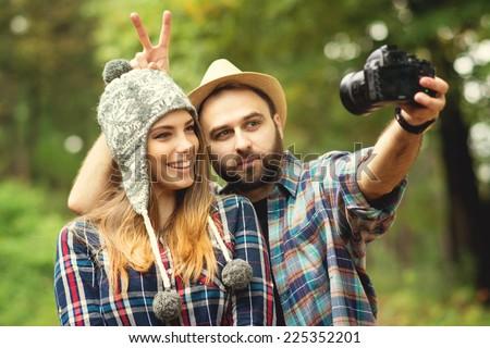 Cute hipster couple with hats taking a selfie with dslr camera in park in autumn. Closeup of young attractive blonde woman and bearded man posing for a self portrait outdoors. 