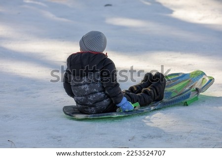 A little boy sitting on a sled on the bottom of a snow hill. A child having fun on the snow.