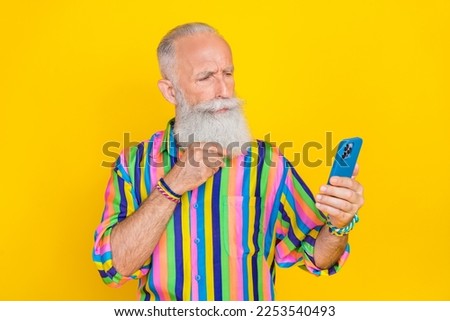 Photo of thoughtful retired man with white beard dressed striped shirt touching beard look at phone isolated on yellow color background