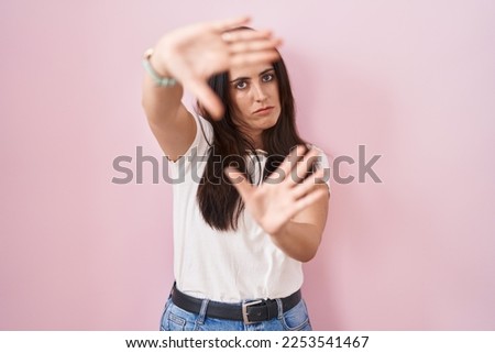 Young brunette woman standing over pink background doing frame using hands palms and fingers, camera perspective 