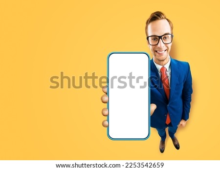 Full body image of businessman in eye glasses, blue suit showing cell phone, mobile smartphone, on yellow background. Comic cartoon funny businessman in eyeglasses with cellphone. Expert recommending.