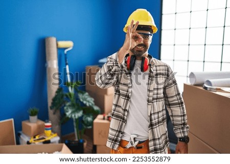 Young hispanic man with beard working at home renovation doing ok gesture with hand smiling, eye looking through fingers with happy face. 