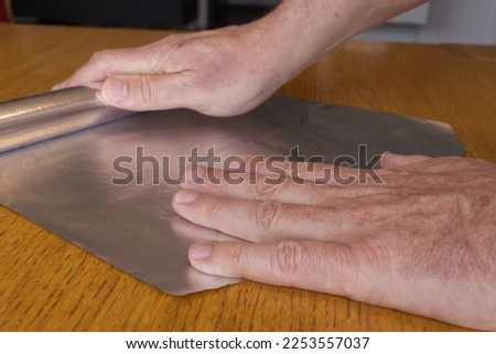 Men hands roll off the aluminum foil for household use on a wooden surface.   Royalty-Free Stock Photo #2253557037