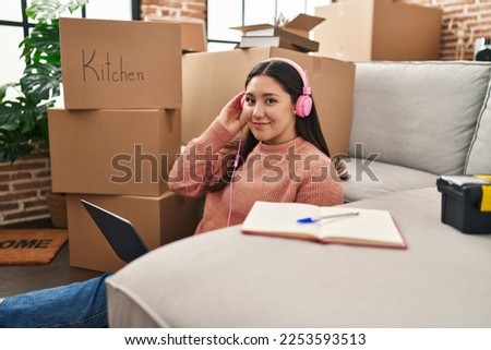 Young hispanic woman using laptop and headphones sitting on floor at new home