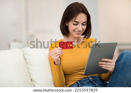 Cute cheerful pretty brunette young lady sitting on couch in living room, holding red credit card and modern digital tablet, making online order or purchasing on Internet, copy space. E-commerce Royalty-Free Stock Photo #2253597839